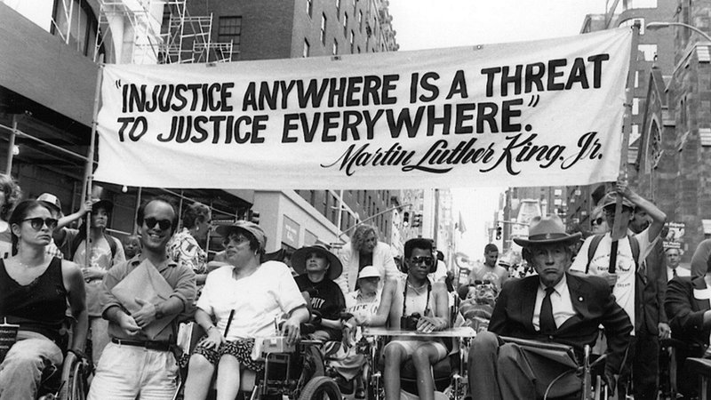 Crowd of disability rights activists under the sign 'Injustice anywhere is a threat to justice everywhere'